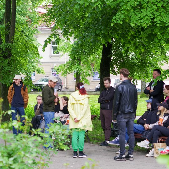 Žmonės stovi ir sėdi po medžiais parke, susirinkę pokalbiui ar poilsiui. Vieni laiko puodelius, kiti bendrauja ar stebi aplinką.