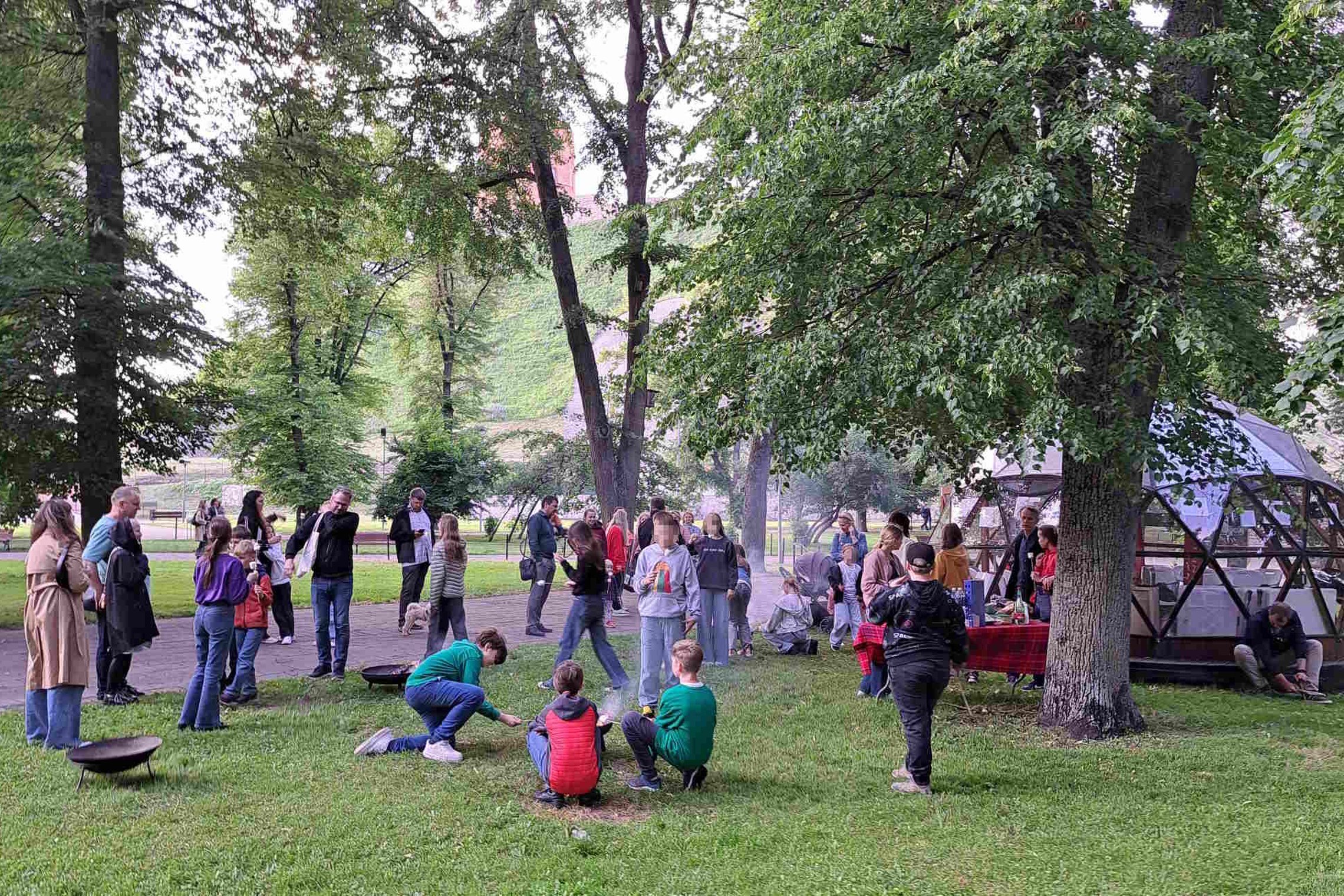 Vaikai ir suaugusieji dalyvauja edukacijoje „Ugnies dirbtuvės“ prie kupolo formos pastato Vilniaus pilių teritorijoje; fone matyti Gedimino pilies bokštas.