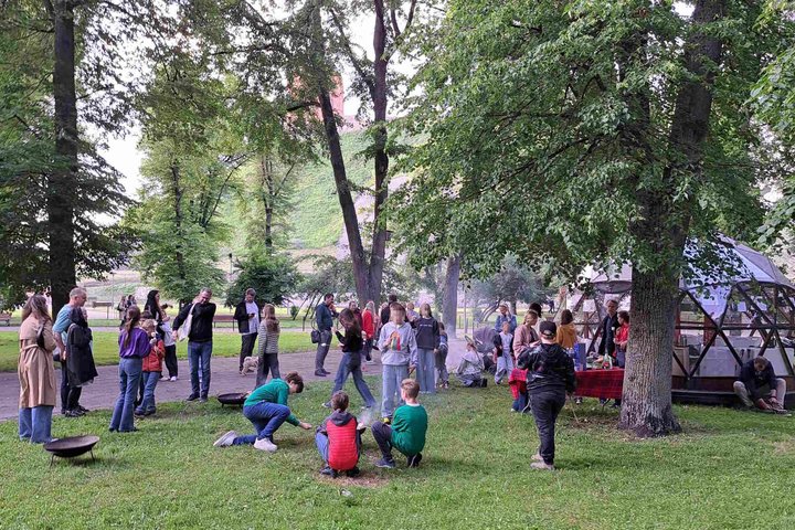 Vaikai ir suaugusieji dalyvauja edukacijoje „Ugnies dirbtuvės“ prie kupolo formos pastato Vilniaus pilių teritorijoje; fone matyti Gedimino pilies bokštas.