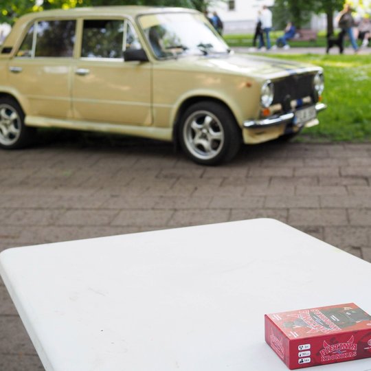 Ant balto stalo stovi stalo žaidimo „Pietinia kronikas“ dėžutė. Fone matyti senas gelsvas automobilis ir žmonės parke.