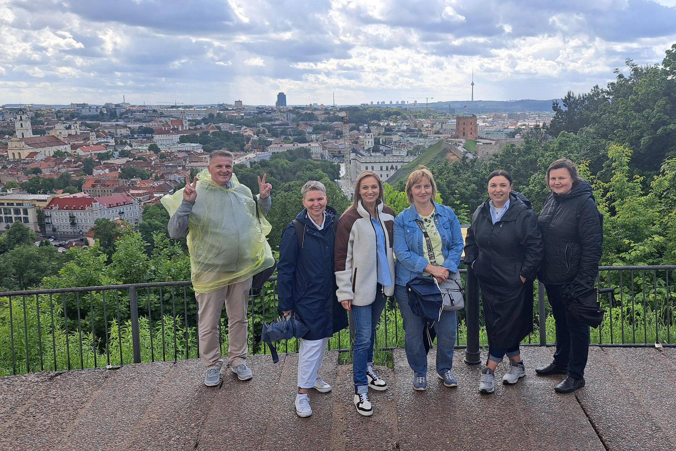 Grupė žmonių po ekskursijos pozuoja ant kalno apžvalgos aikštelės su vaizdu į Vilniaus senamiestį ir Gedimino pilies bokštą; kai kurie dėvi lietpalčius, fone matyti debesuotas dangus