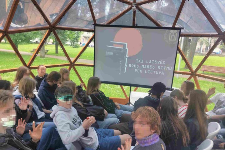 Moksleiviai sėdi kupolo formos erdvėje ir dalyvauja edukacijoje „Iki laisvės Roko maršo ritmu per Lietuvą“, rodomoje projektoriaus ekrane.