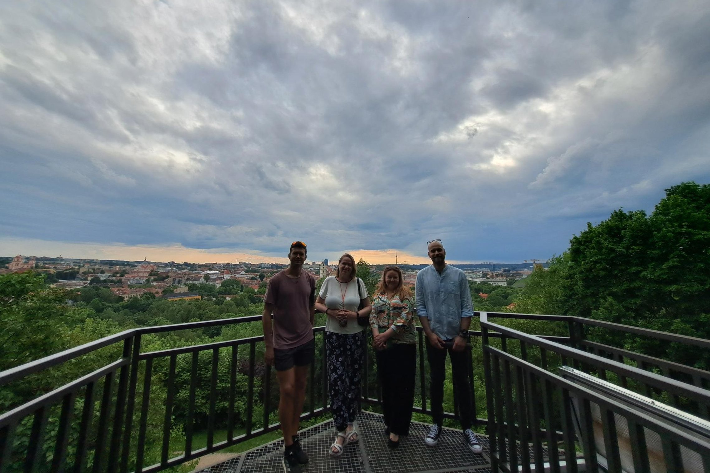 Keturi ekskursijos dalyviai stovi apžvalgos aikštelėje su plieniniais turėklais, už jų atsiveria Vilniaus senamiesčio panorama. Dangų dengia dramatiški debesys.