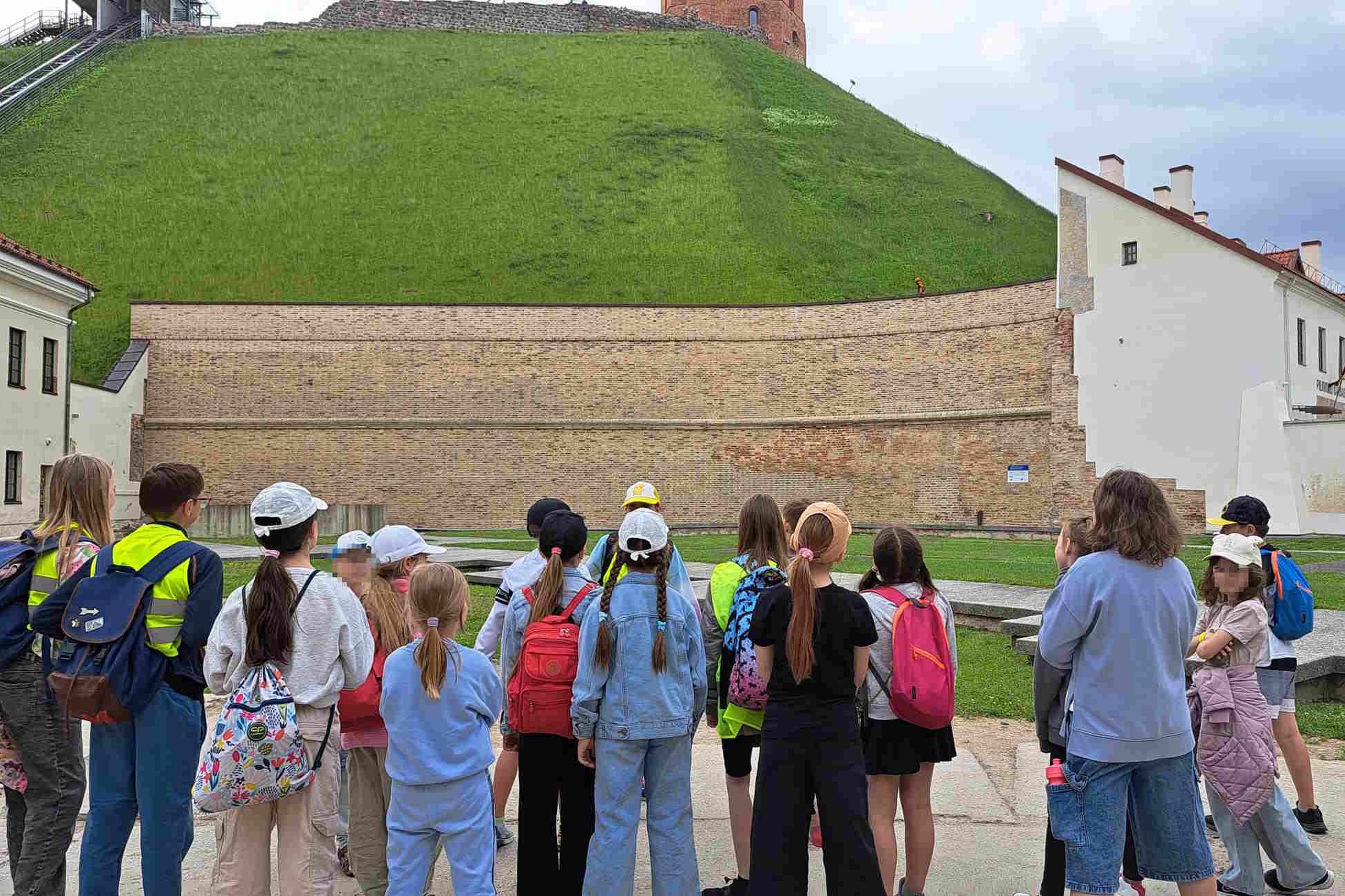 Vaikų grupė stovi priešais Gedimino kalną Vilniuje, klausosi edukacijos apie Vilniaus pilis; fone matyti mūrinė kalno atraminė siena ir Gedimino bokštas viršuje.
