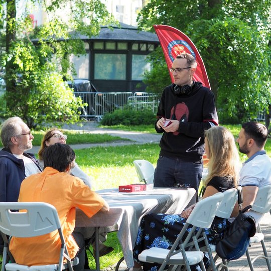 Vyras stovi prie stalo parke ir kalba su aplink sėdinčia grupe žmonių. Už jo plėvesuoja raudona „Vilniaus pilių“ vėliava, fone matyti medžiai ir pastatai.