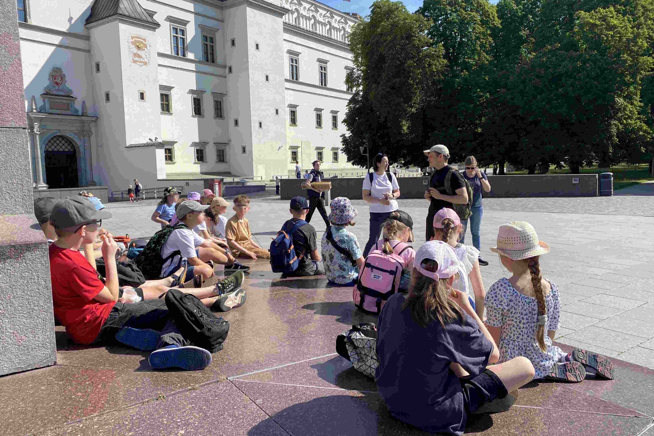 Vaikai su kuprinėmis ir kepurėlėmis sėdi ant grindinio Vilniaus Katedros aikštėje, klausosi edukatoriaus prie Valdovų rūmų fono.