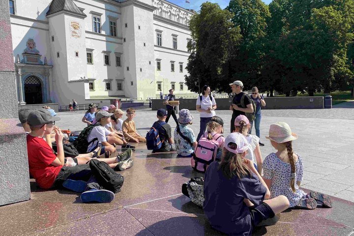 Vaikai su kuprinėmis ir kepurėlėmis sėdi ant grindinio Vilniaus Katedros aikštėje, klausosi edukatoriaus prie Valdovų rūmų fono.