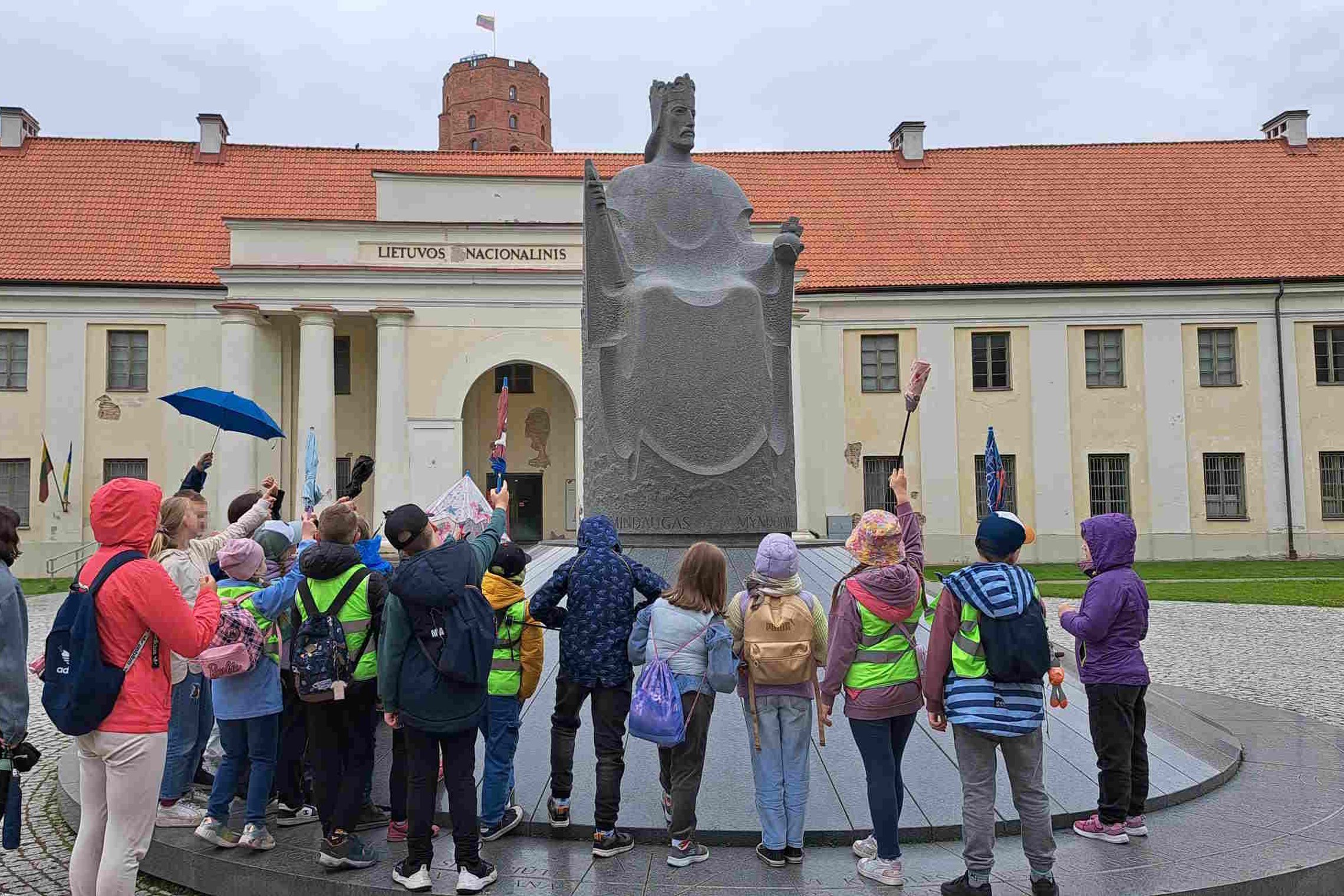 Vaikų grupė su vadovais stovi prie Mindaugo paminklo Vilniaus pilių teritorijoje, fone – Lietuvos nacionalinis muziejus ir Gedimino pilies bokštas.