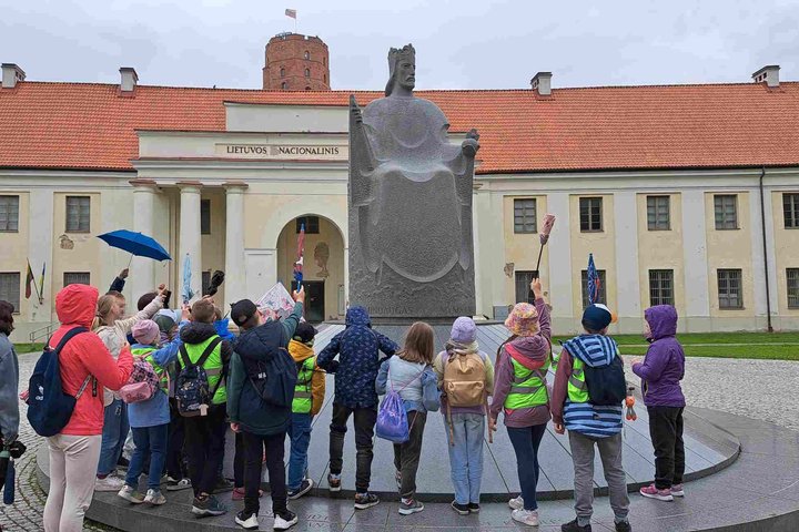 Vaikų grupė su vadovais stovi prie Mindaugo paminklo Vilniaus pilių teritorijoje, fone – Lietuvos nacionalinis muziejus ir Gedimino pilies bokštas.