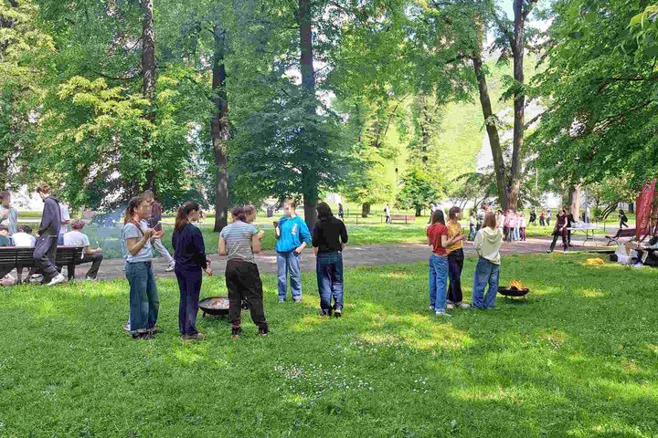 Grupės paauglių stovi prie Vilniaus pilių kupolo degančių laužaviečių, dalis jų valgo, kiti kalbasi; fone matyti kiti žmonės, suolai, kupolas ir medžiai.