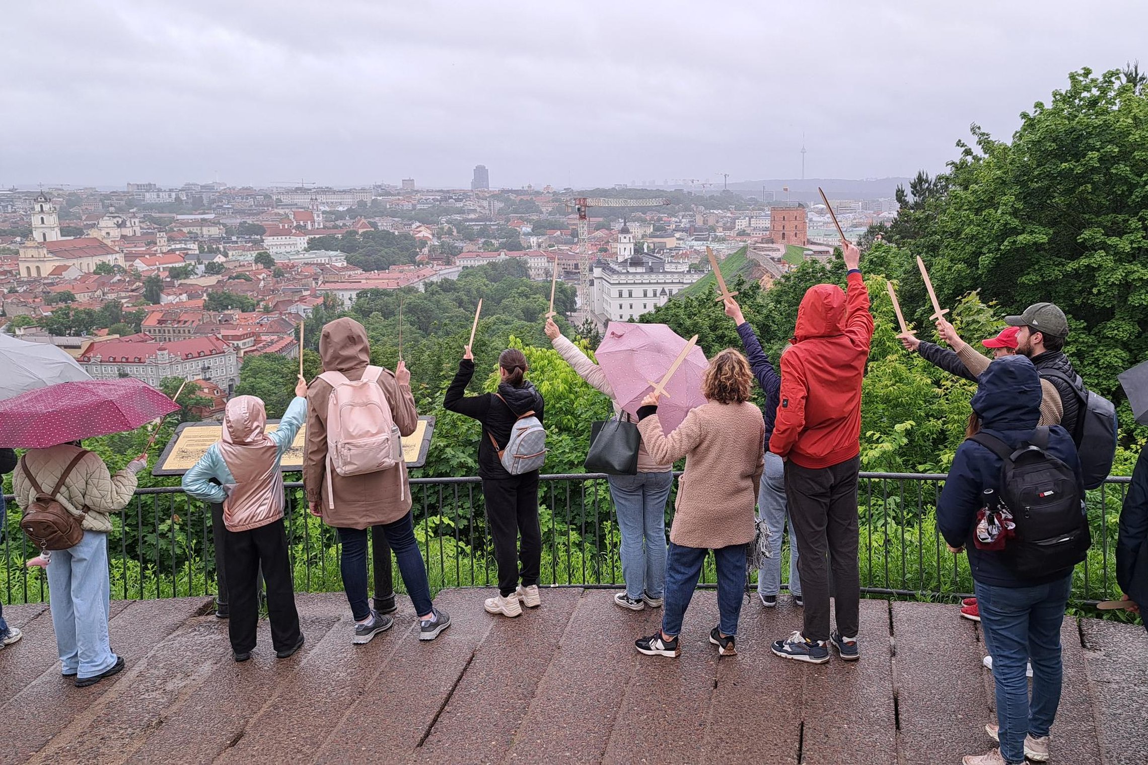 Grupė žmonių su skėčiais ir lietpalčiais stovi ant apžvalgos aikštelės lietingą dieną ir rodo medinėmis rodyklėmis į Vilniaus senamiestį bei Gedimino pilies bokštą.