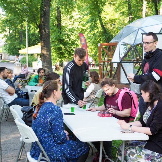 Žmonės sėdi prie stalų parke ir kalbasi, vienas vyras pila gėrimą į plastikinius puodelius. Fone matyti kupolas, raudona „Vilniaus pilių“ vėliava ir stalo žaidimo dėžutė ant stalo.