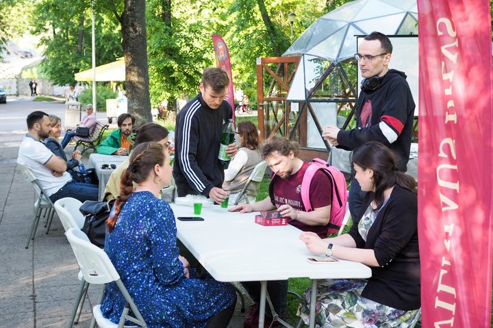 Žmonės sėdi prie stalų parke ir kalbasi, vienas vyras pila gėrimą į plastikinius puodelius. Fone matyti kupolas, raudona „Vilniaus pilių“ vėliava ir stalo žaidimo dėžutė ant stalo.