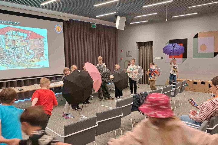 Vaikai edukacijoje apie vandens malūną TELIA patalpose stovi puslankiu, laikydami įvairiaspalvius skėčius, priešais rodomą skaidrę su malūno pjūvio iliustracija.