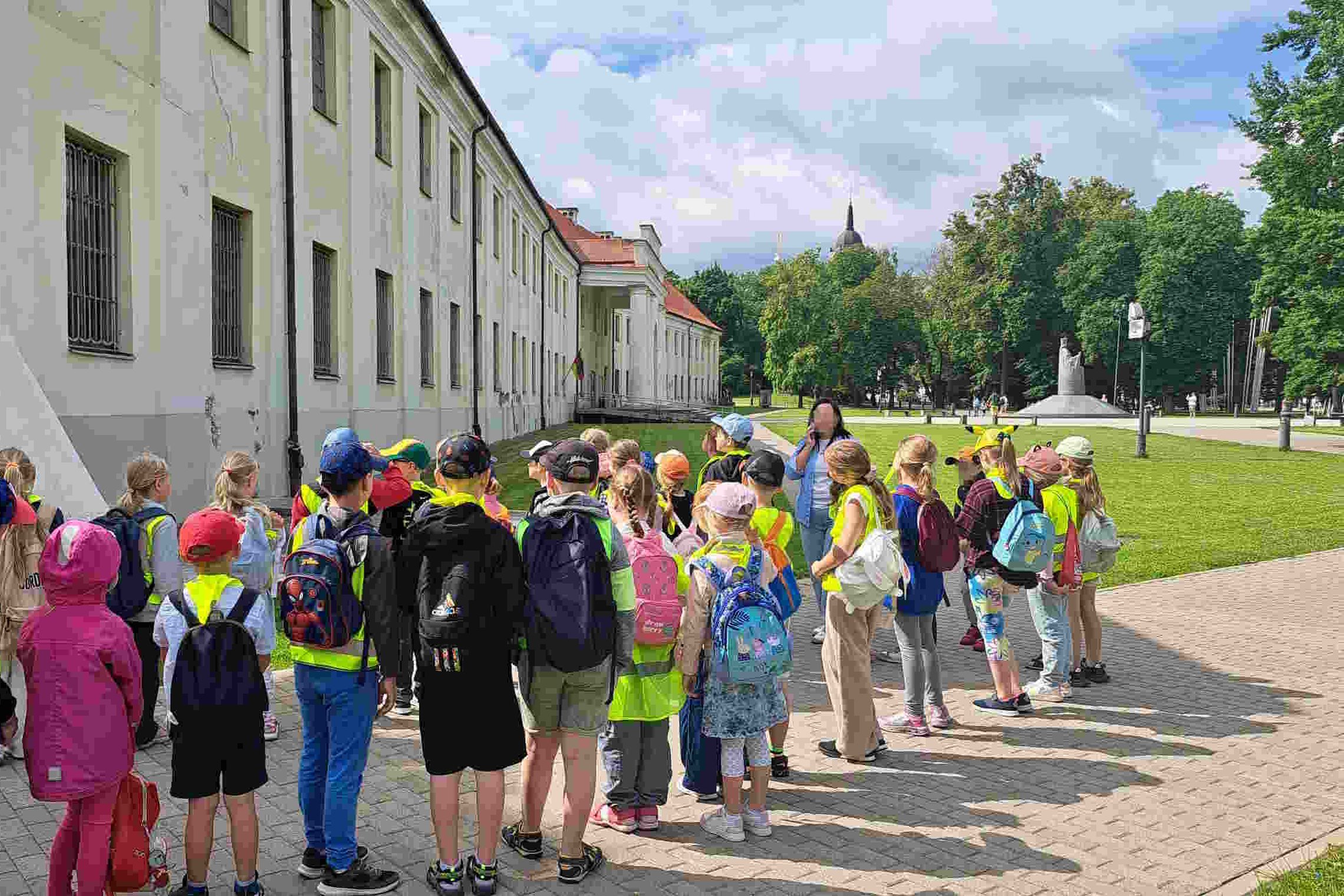 Grupė vaikų su ryškiaspalvėmis liemenėmis ir kuprinėmis stovi prie balto pastato Vilniaus centre, klausosi edukatoriaus pasakojimo; fone matyti žaliuojantis parkas ir paminklas Mindaugo.