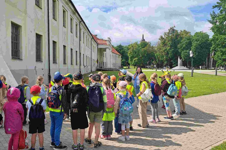 Grupė vaikų su ryškiaspalvėmis liemenėmis ir kuprinėmis stovi prie balto pastato Vilniaus centre, klausosi edukatoriaus pasakojimo; fone matyti žaliuojantis parkas ir paminklas Mindaugo.