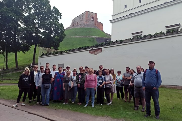 Ekskursijos dalyviai pozuoja grupinei nuotraukai prie Senojo arsenalo sienos Vilniuje, Gedimino kalnas su pilimi matyti fone. Grupėje stovi įvairaus amžiaus žmonės, aplink žaliuoja žolė ir medžiai.