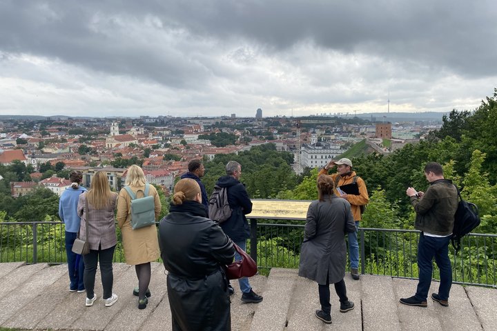 Ekskursijos dalyviai stovi apžvalgos aikštelėje, žvelgdami į Vilniaus senamiestį; priekyje – gidas, aiškinantis apie miesto istoriją, tolumoje matyti Gedimino pilies bokštas ir Vilniaus televizijos bokštas.
