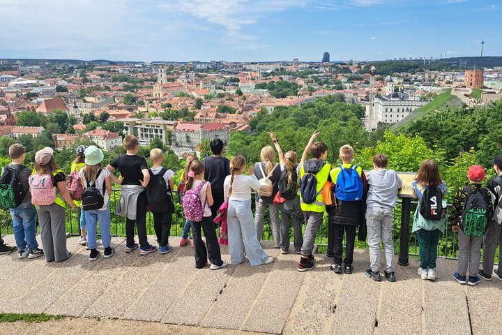 Trečiokų grupė iš Žėručio progimnazijos stovi prie Kalnų parko apžvalgos aikštelės ir žvelgia į Vilniaus senamiestį; tolumoje matyti Gedimino pilies bokštas, bažnyčių bokštai ir miesto stogai.