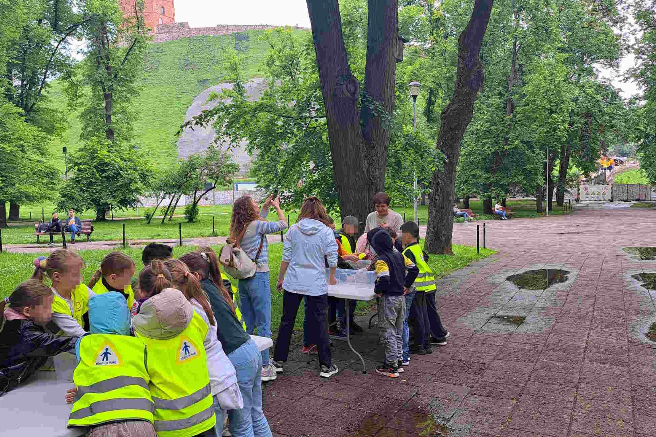 Vaikai su geltonomis liemenėmis dalyvauja edukacijoje lauke prie stalo, fone matyti žalias Gedimino kalnas su pilies bokštu.
