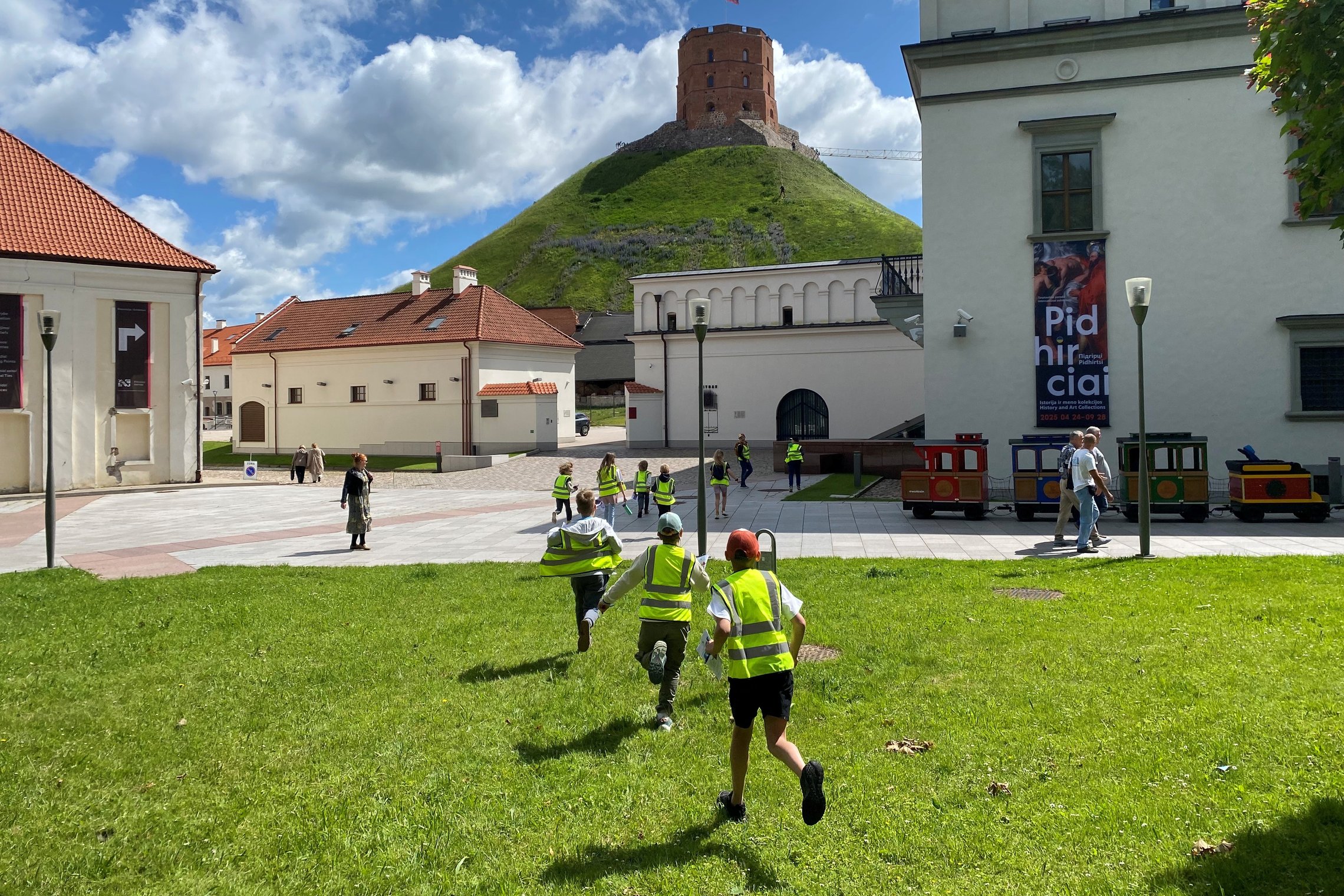 Vaikai bėga per Vilniaus pilių muziejaus kiemą link Gedimino kalno, fone matyti Gedimino pilies bokštas.