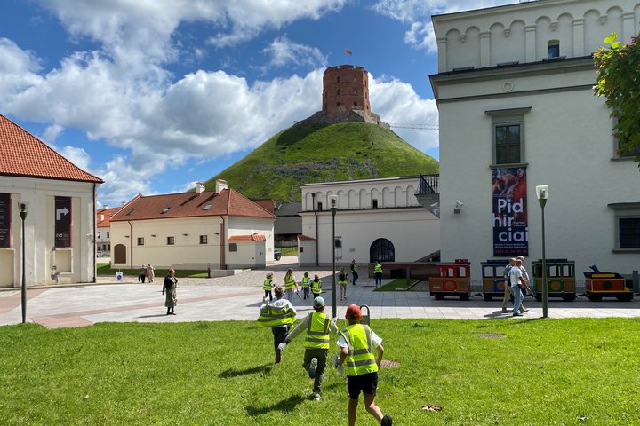 Vaikai bėga per Vilniaus pilių muziejaus kiemą link Gedimino kalno, fone matyti Gedimino pilies bokštas.
