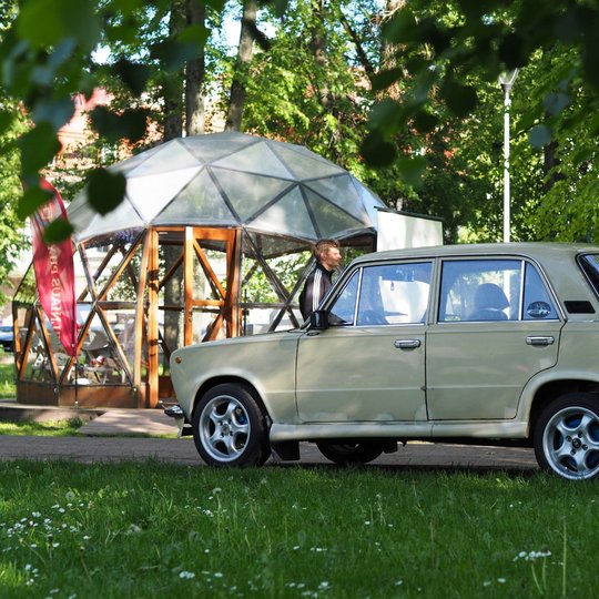 Parke stovi senas gelsvas automobilis, šalia jo – vyras atidarytomis durelėmis. Fone matyti stiklinis kupolas ir raudona „Vilniaus pilių“ vėliava.