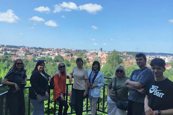 Grupė žmonių stovi Altanos kalno apžvalgos aikštelėje Vilniaus pilių teritorijoje. Fone – platus Vilniaus senamiesčio vaizdas su raudonais stogais, bažnyčių bokštais ir žaliomis medžių viršūnėmis. Dangus giedras, su keliomis baltais debesimis.