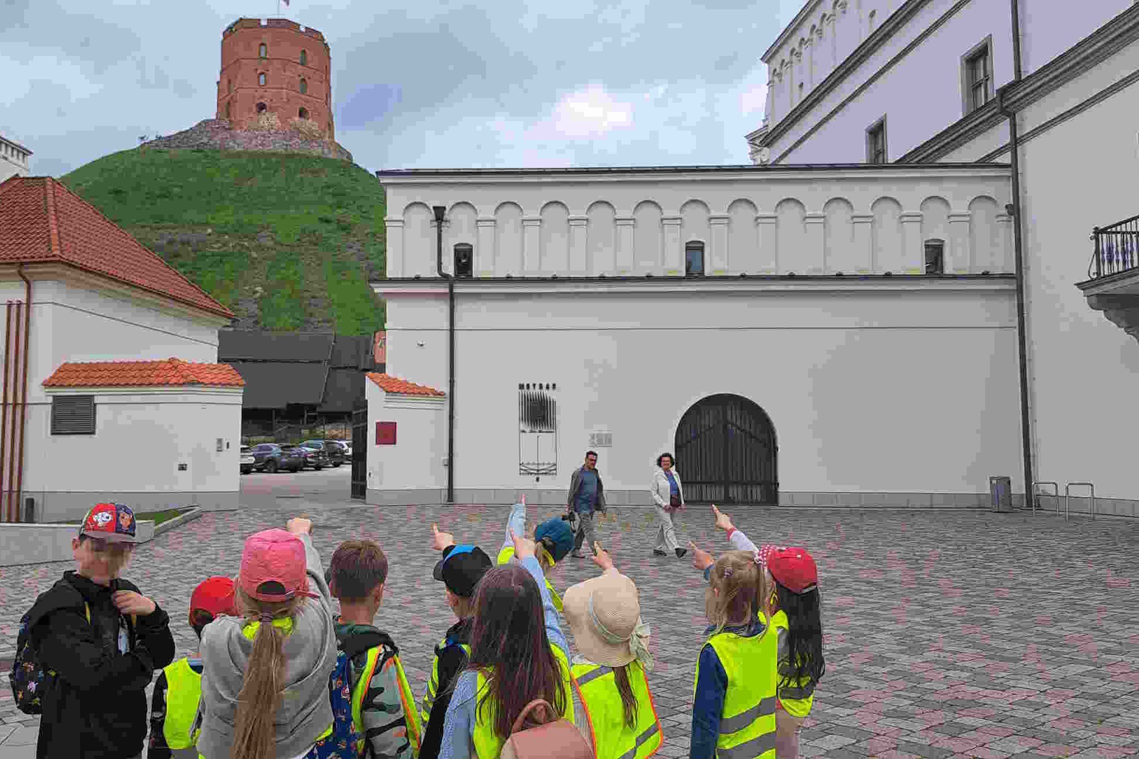 Vaikai su ryškiaspalvėmis liemenėmis ir kepuraitėmis, rodo rankomis į Gedimino pilies bokštą, esantį kalno viršuje. Fone – Vilniaus pilių komplekso pastatai.