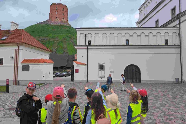 Vaikai su ryškiaspalvėmis liemenėmis ir kepuraitėmis, rodo rankomis į Gedimino pilies bokštą, esantį kalno viršuje. Fone – Vilniaus pilių komplekso pastatai.