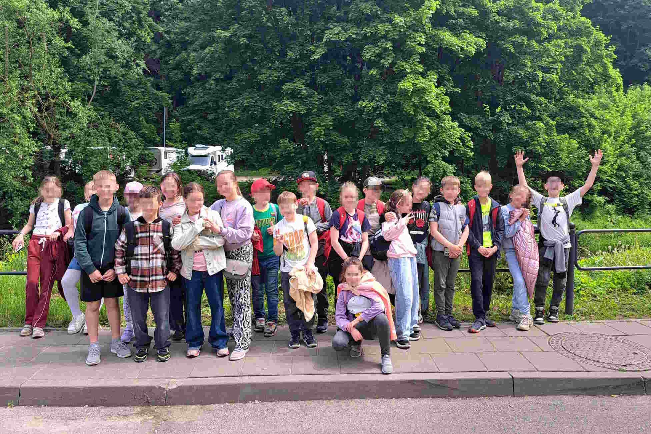Vilniaus Maironio progimnazijos trečiokų klasė stovi prie Kalnų parko tako žaliuojančių medžių fone; vaikai šypsosi, dalis laiko užkandžius ar rankas iškėlę į viršų.