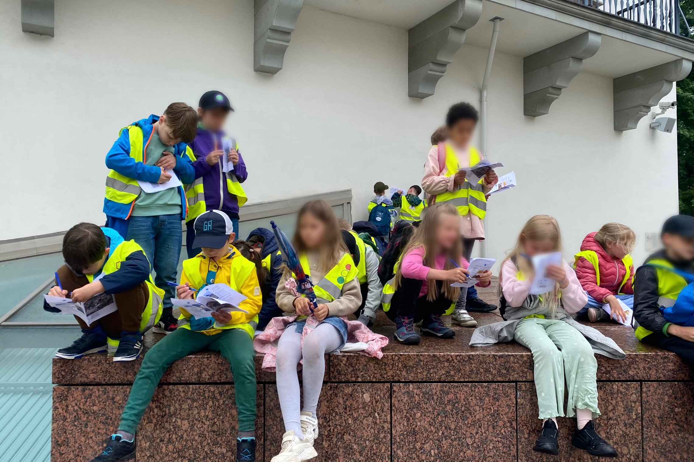 Edukacinėje veikloje dalyvaujantys pradinių klasių mokiniai prie muziejaus pastato atlieka užduotis iš edukacinių knygelių