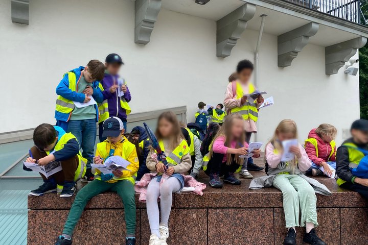 Edukacinėje veikloje dalyvaujantys pradinių klasių mokiniai prie muziejaus pastato atlieka užduotis iš edukacinių knygelių