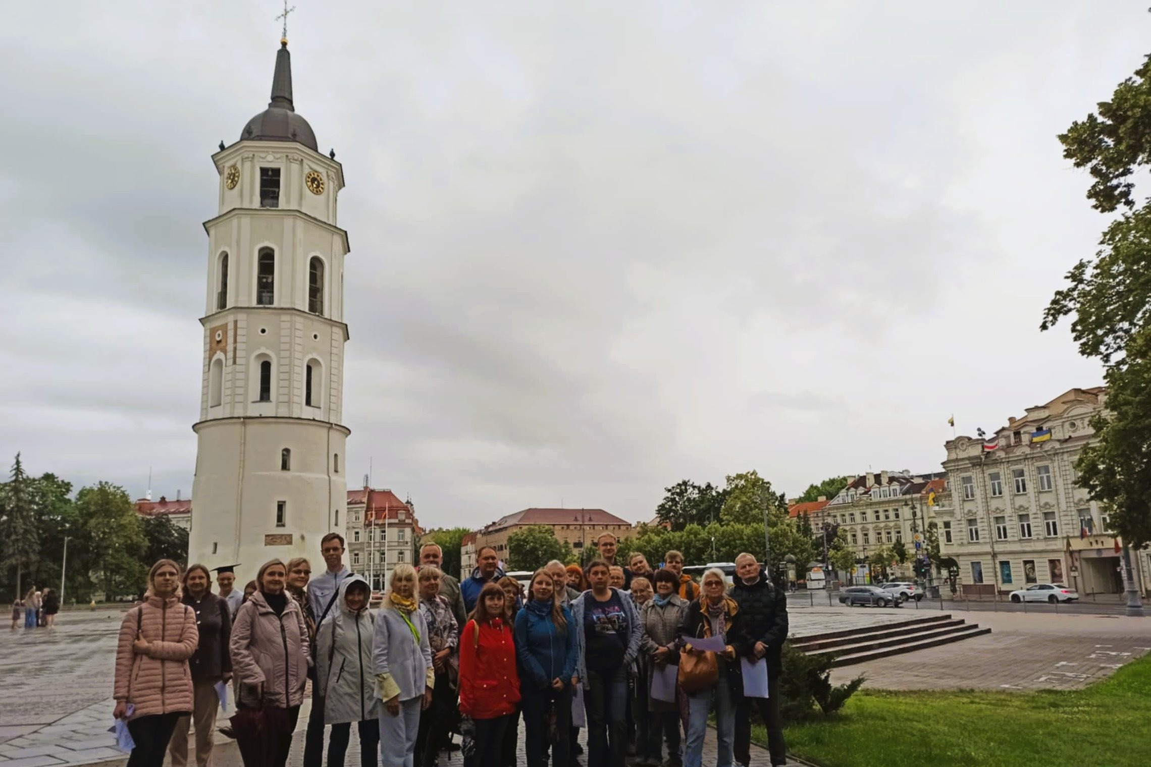 Nuotraukoje grupė žmonių pozuoja prie Vilniaus Katedros varpinės. Kai kurie žmonės rankose laiko popieriaus lapus - galimai tai ekskursijos vaizdinė medžiaga.