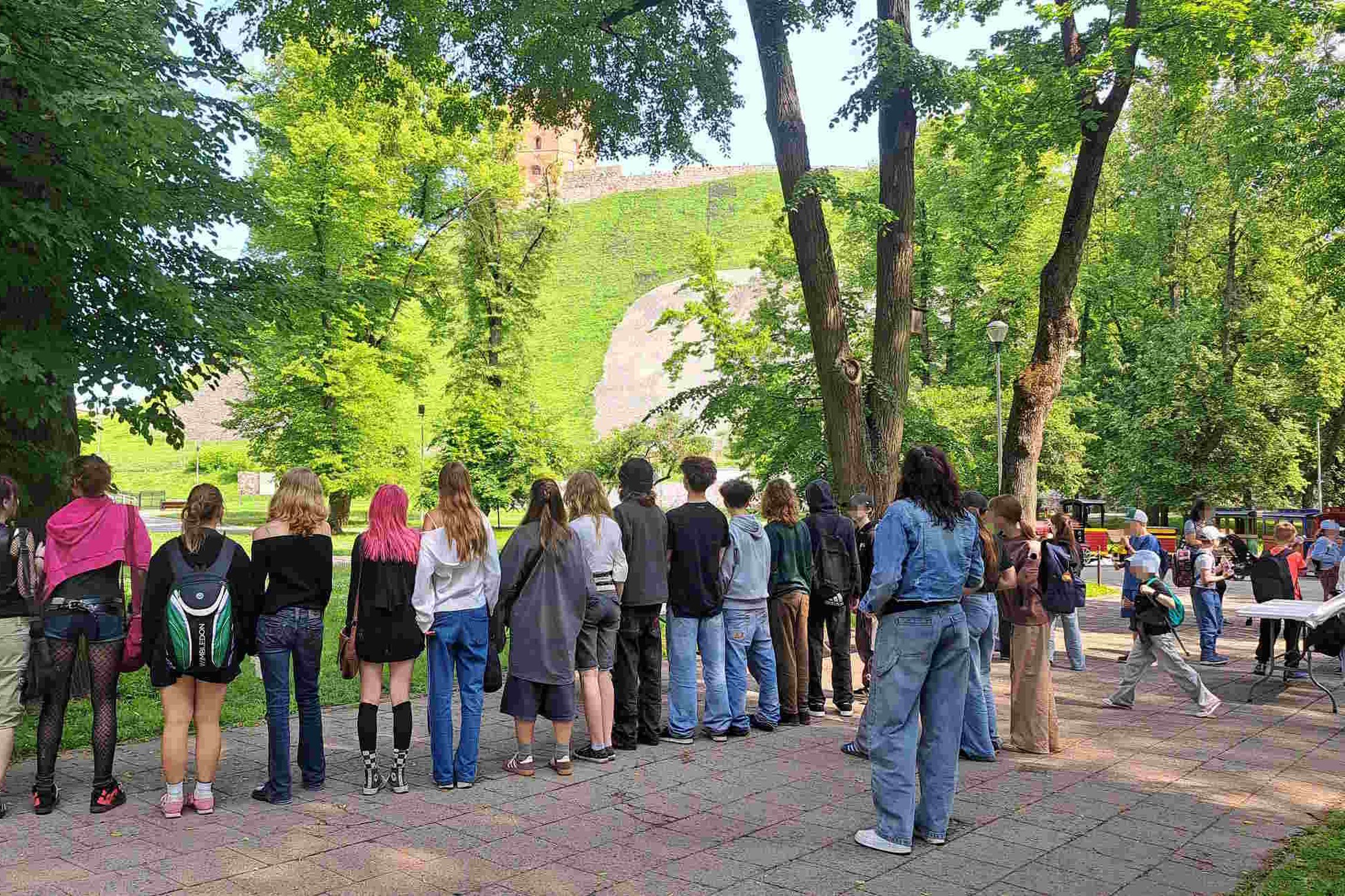 Mokiniai stovi nugara į kamerą Kalnų parke, priešais matyti žaliuojantis Gedimino kalnas ir bokštas; dešinėje – žmonės prie staliukų, vyksta renginys.