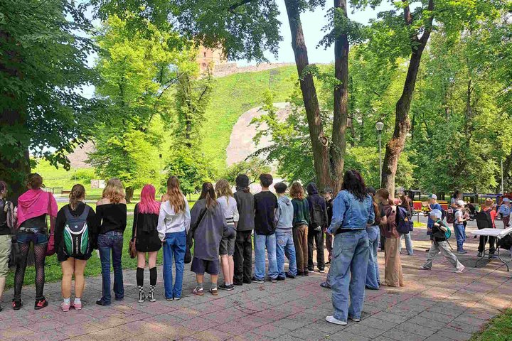 Mokiniai stovi nugara į kamerą Kalnų parke, priešais matyti žaliuojantis Gedimino kalnas ir bokštas; dešinėje – žmonės prie staliukų, vyksta renginys.