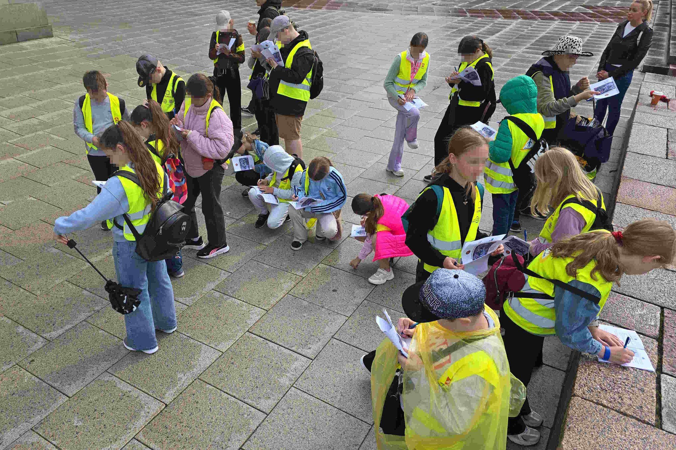 **Alt tekstas:** Vaikai su ryškiais geltonais liemenėmis stovi ir sėdi prie lauko akmeninių grindinių, rašo užduočių lapuose ir tyrinėja aplinką per edukaciją „Žemutinės pilies paslaptys“.