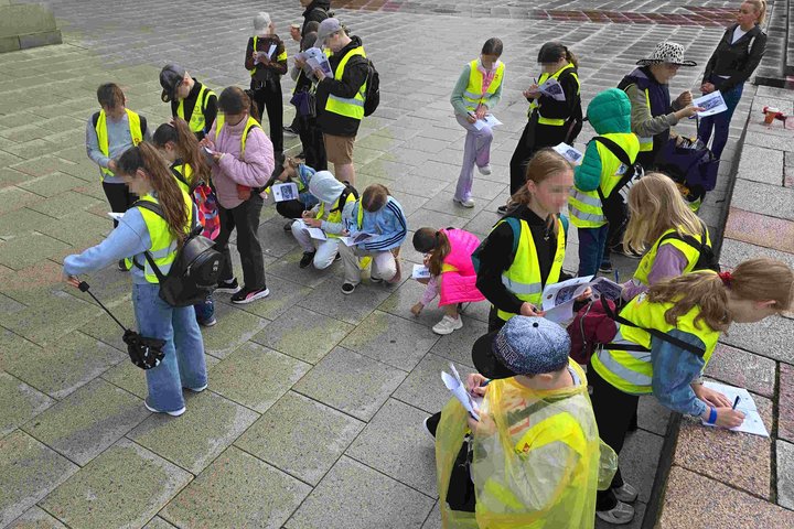 **Alt tekstas:** Vaikai su ryškiais geltonais liemenėmis stovi ir sėdi prie lauko akmeninių grindinių, rašo užduočių lapuose ir tyrinėja aplinką per edukaciją „Žemutinės pilies paslaptys“.