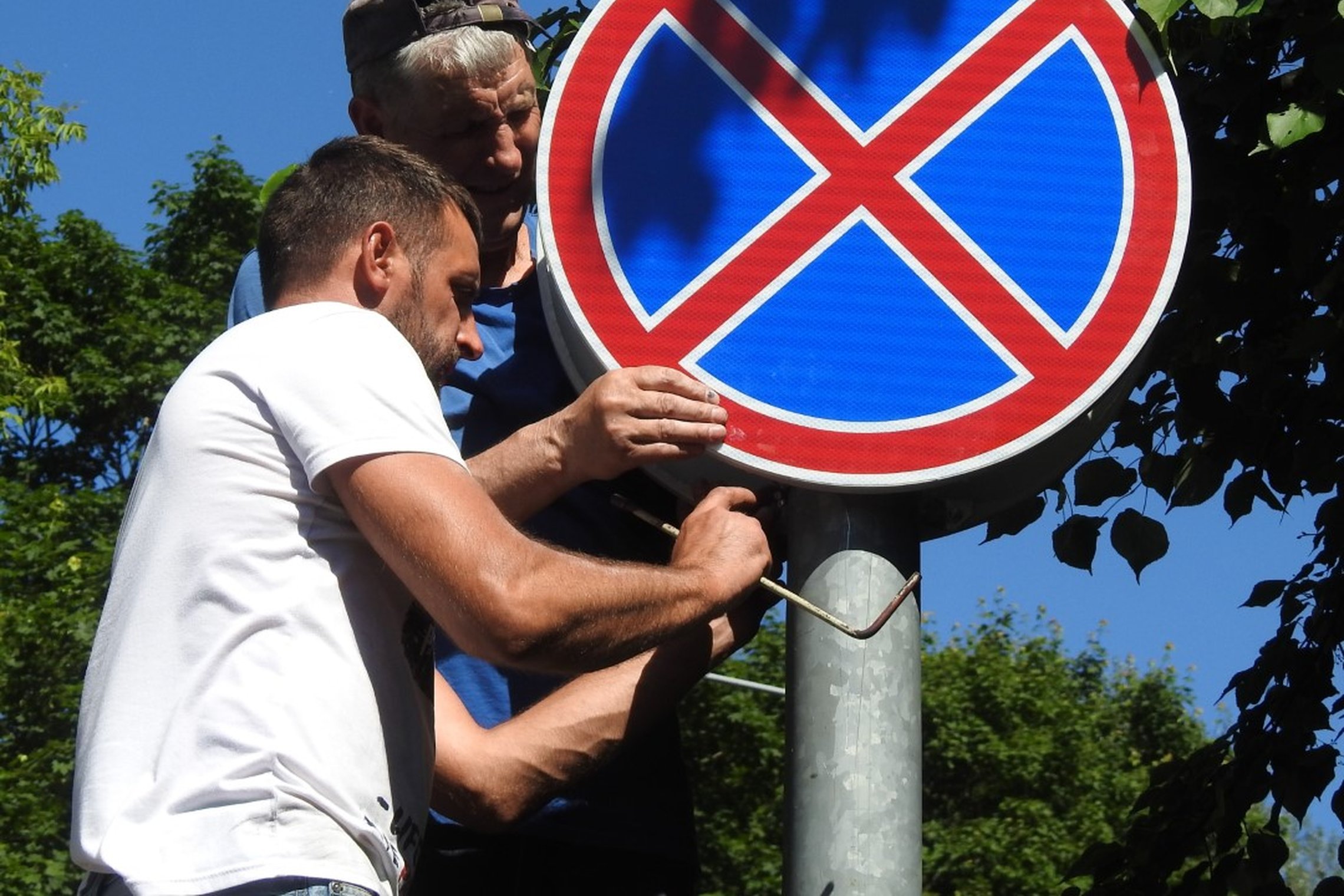 Nuotraukoje du vyrai stato kelio ženklą. Vyrai apsirengę trumpų rankovių maikutėm. Aplinkui daug tankių medžių su lapais. Tai vasaros diena.