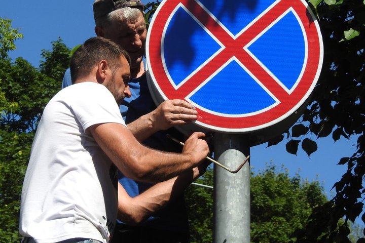 Nuotraukoje du vyrai stato kelio ženklą. Vyrai apsirengę trumpų rankovių maikutėm. Aplinkui daug tankių medžių su lapais. Tai vasaros diena.
