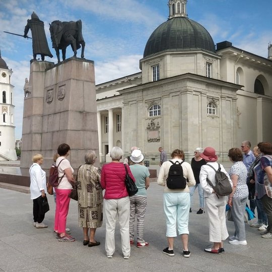 Grupė ekskursijos dalyvių būreliu sustoję prie Gedimino paminklo Katedros aikštėje.
