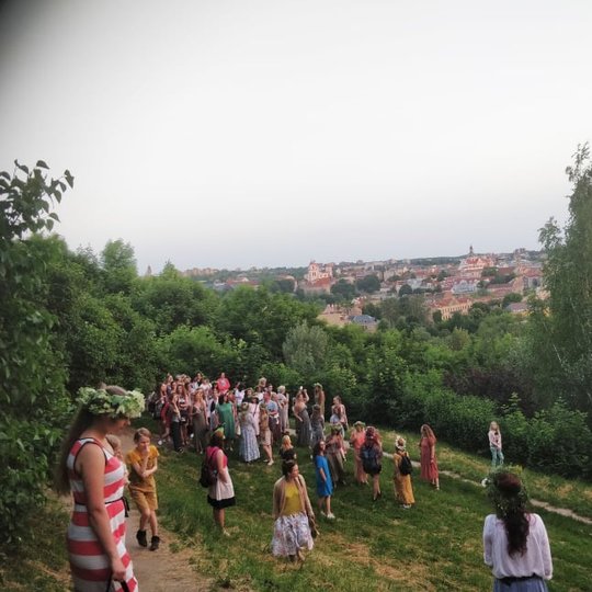 Vakarėjanti diena ir daug moterų, pasipuošusių vasarinių gėlių vainikais, stovi ant Altanos kalno šlaito ir žiūri į nuostabų vaizdą - Vilniaus miestas apšvietas besileidžiančios saulės yra po kojomis.