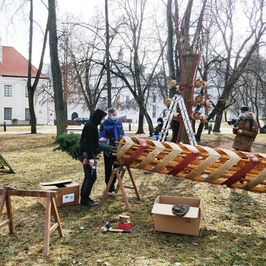 Mindaugo paminklo skvere dar neatsigavusi po žiemos veja. Ant vejos keli žmonės stato milžinišką verbą. Verbų skulptūros šiemet pasirinktos kaip Pavasario lygiadienį įprasminantis bei gyvybės medį atspindintis simbolis.