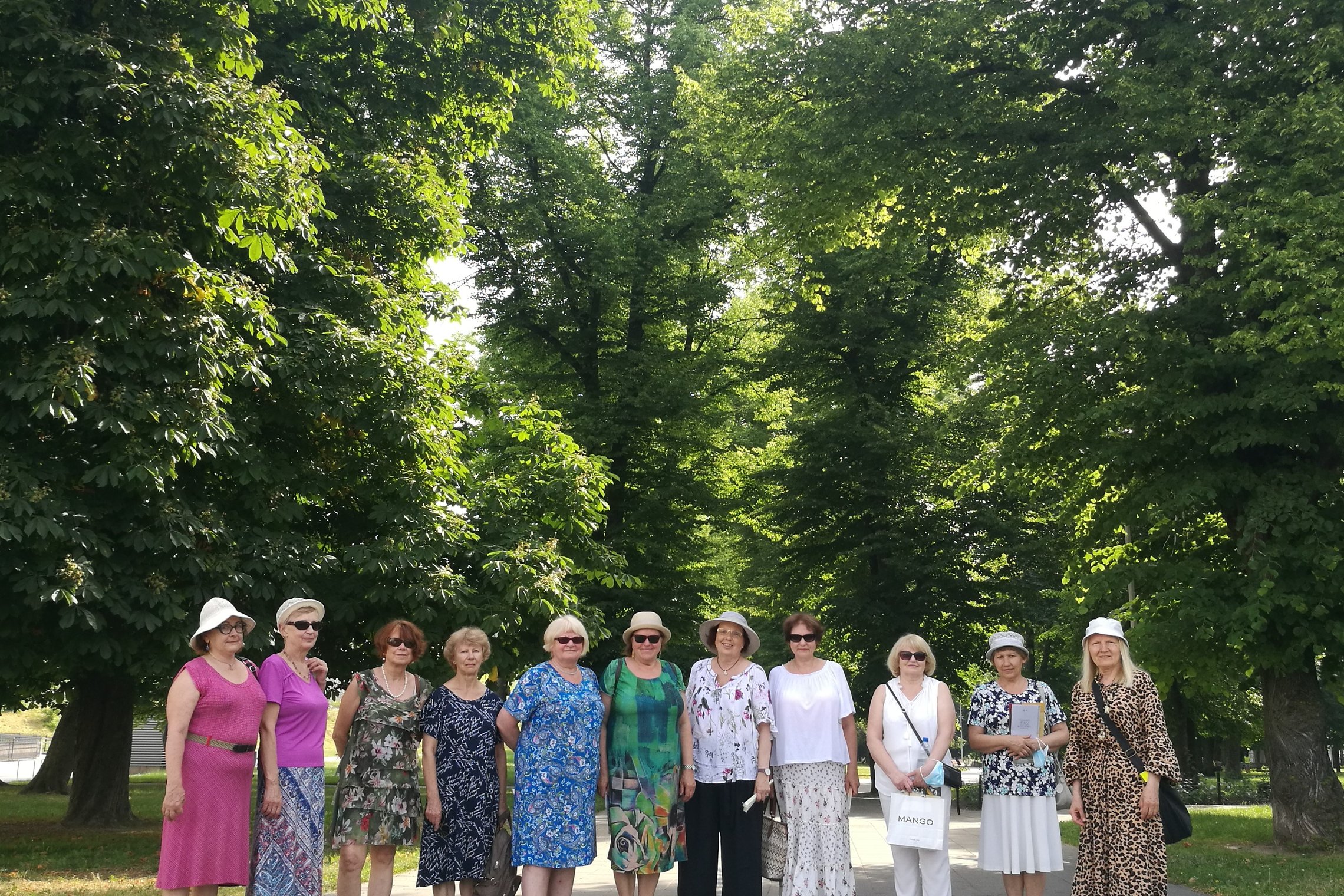 Parko alėjoje stovi grupė moterų. Jos vasarinėmis suknelėmis ir šviesiomis kepuraitėmis. Už jų nugaros yra seni medžiai. Saulėta graži diena.
