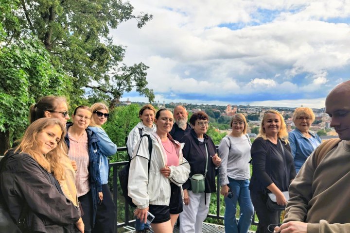 Grupė žmonių stovi Altanos kalno apžvalgos aikštelėje, Vilniuje, su vaizdu į miesto panoramą fone. Dalyviai šypsosi ir žvelgia į kamerą, kai kurie atsirėmę į turėklą. Už jų matyti žali medžiai ir tolumoje – Vilniaus stogai, bažnyčių bokštai, Gedimino pili