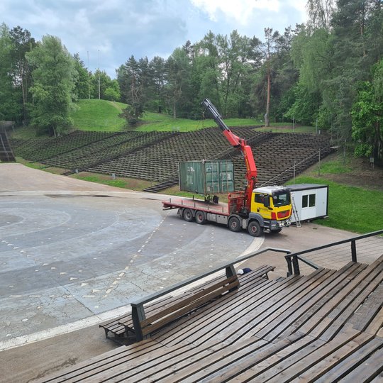 Dainų slėnio estrada po remonto. Nauji mediniai suolai. Didelė mašina su kranu kelią statybininkų psakutinę būdelę - remontas baigia, statybininkai palieka suremontuotą estradą.