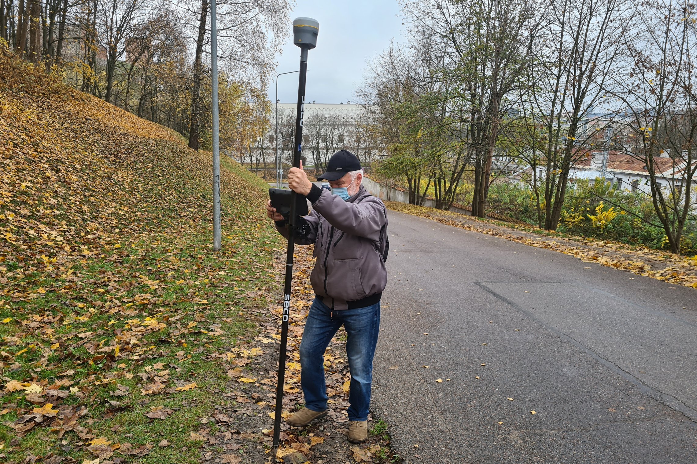 Nuotraukoje žilas žmogus, kuris yra su juodos spalvos kepure, pilkos spalvos striuke, mėlynais džinsais ir smėlio spalvos batais. Jis taip pat užsidėjęs šviesiai mėlynos spalvos veido kaukę. Vyras rankose laiko juodos spalvos matavimo prietaisą. Už jo deš