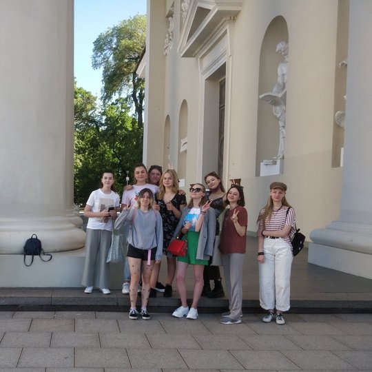 Jaunimo grupelė prie Vilniaus katedros kolonos ir pačios Katedros. Mokiniai apsirengę vasariškai, yra šilta vasaros diena.