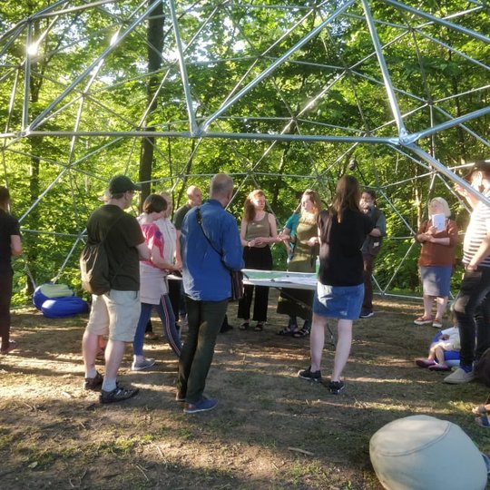 Nemažai susidomėjusių žmonių. Stovi gamtoje, tačiau veja be žolės. Aplinkui miškas, daug medžių su lapais. Per vidurį stovi stalas, o aplinkui keletas sėdmaišių. Visi žmonės stovi po apvaliu iš metalinių konstrukcijų padarytu stogu.