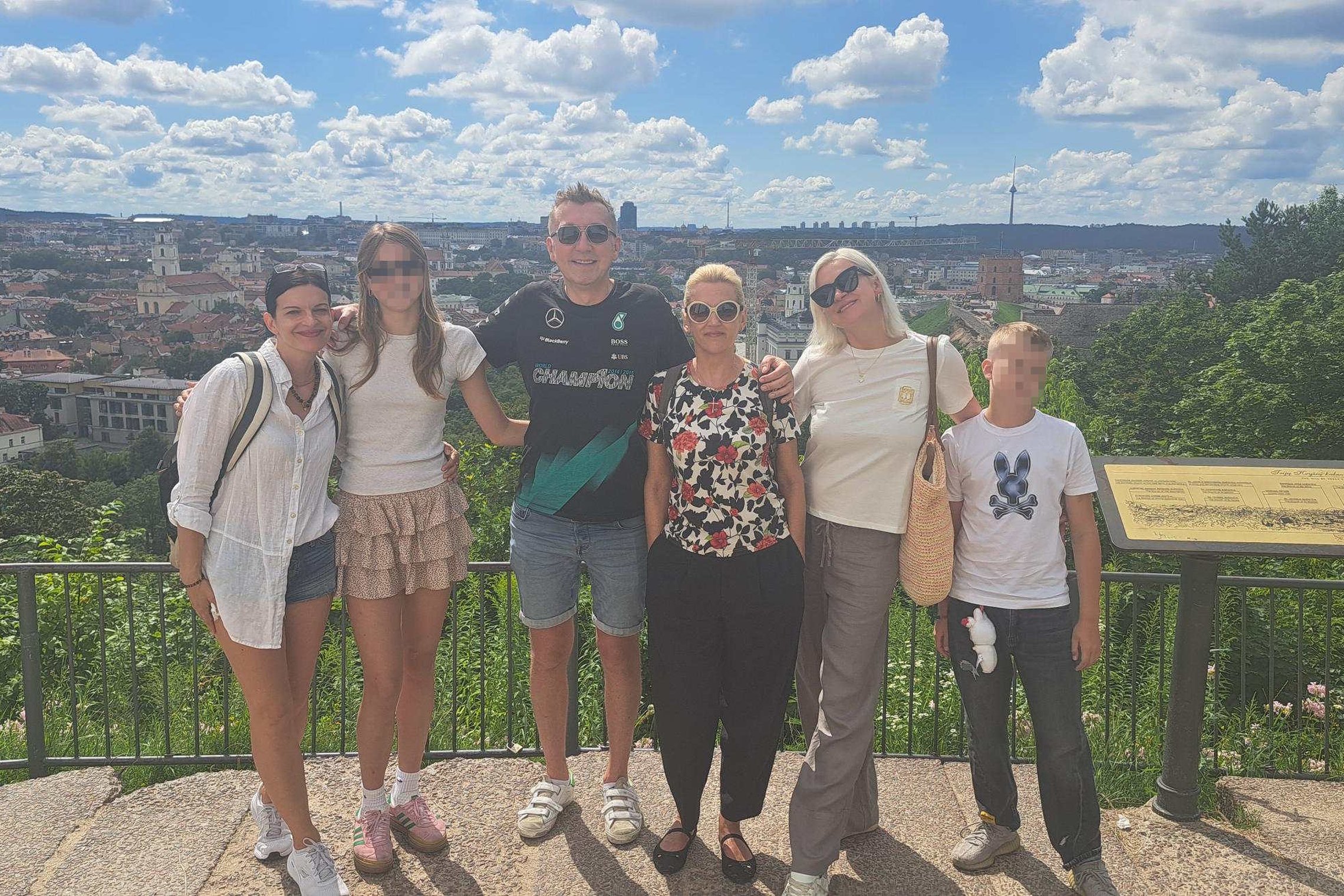 Grupė žmonių stovi ant apžvalgos aikštelės su vaizdu į Vilniaus senamiestį; fone matyti Gedimino pilies bokštas, bažnyčių bokštai ir Televizijos bokštas. Oras saulėtas, dangus giedras su baltais debesimis.